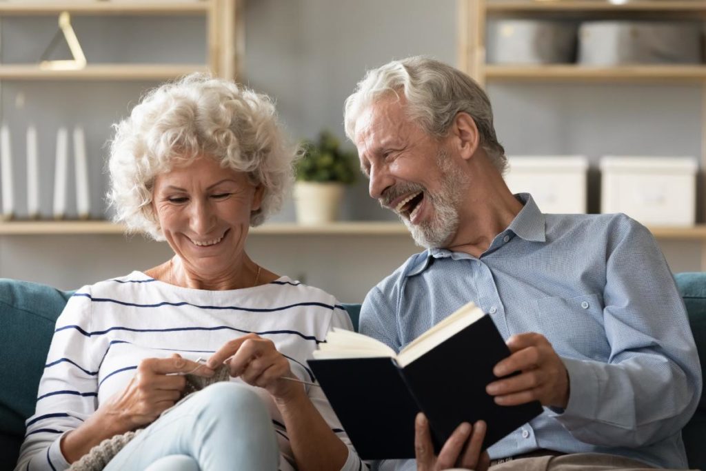 Senior couple on couch in assisted living facility, reading and laughing